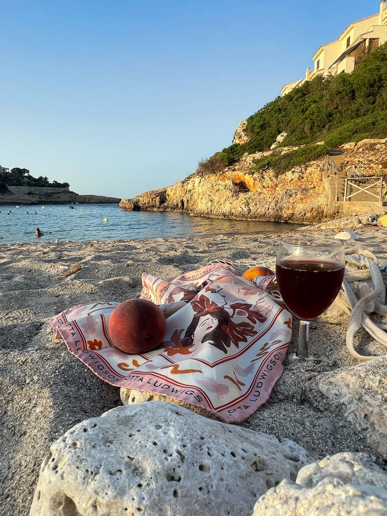 A serene beach scene shows a glass of red wine on a colorful scarf laying on sandy ground. A peach and a peeled orange rest nearby. In the background, clear blue water extends towards rocky cliffs and a few distant swimmers. Sunlight casts a warm glow.