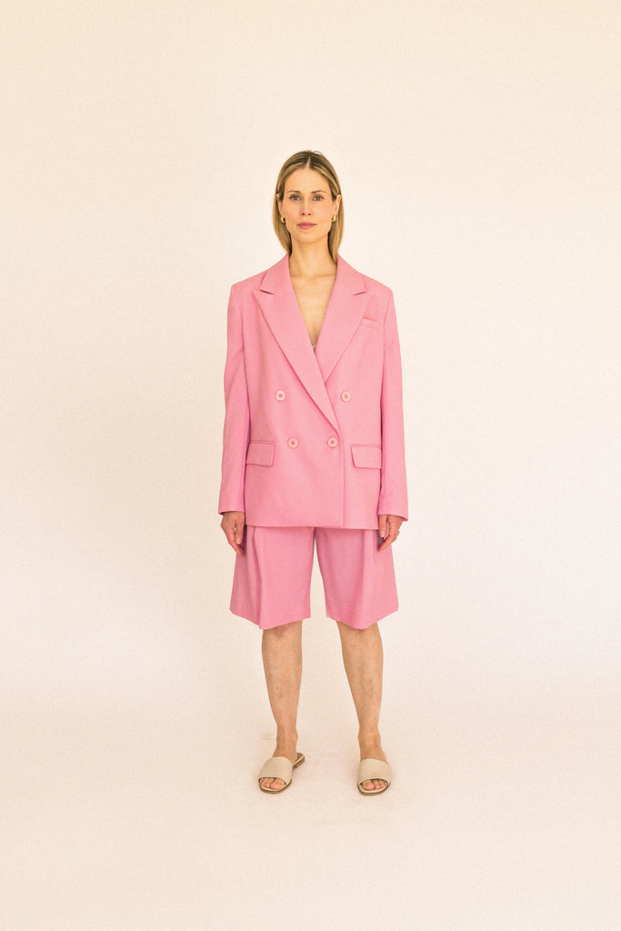 A person with long blond hair stands against a plain white background, wearing the ALVA Blazer in Candy Pink and matching pants. 