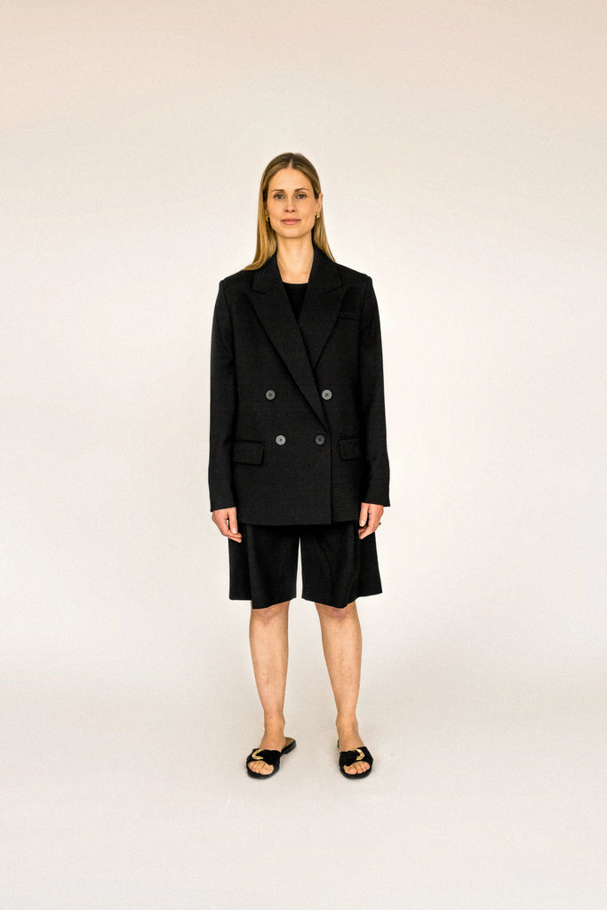 A person with long blond hair stands against a plain white background, wearing the ALVA Blazer in Tuxedo Black and matching shorts.
