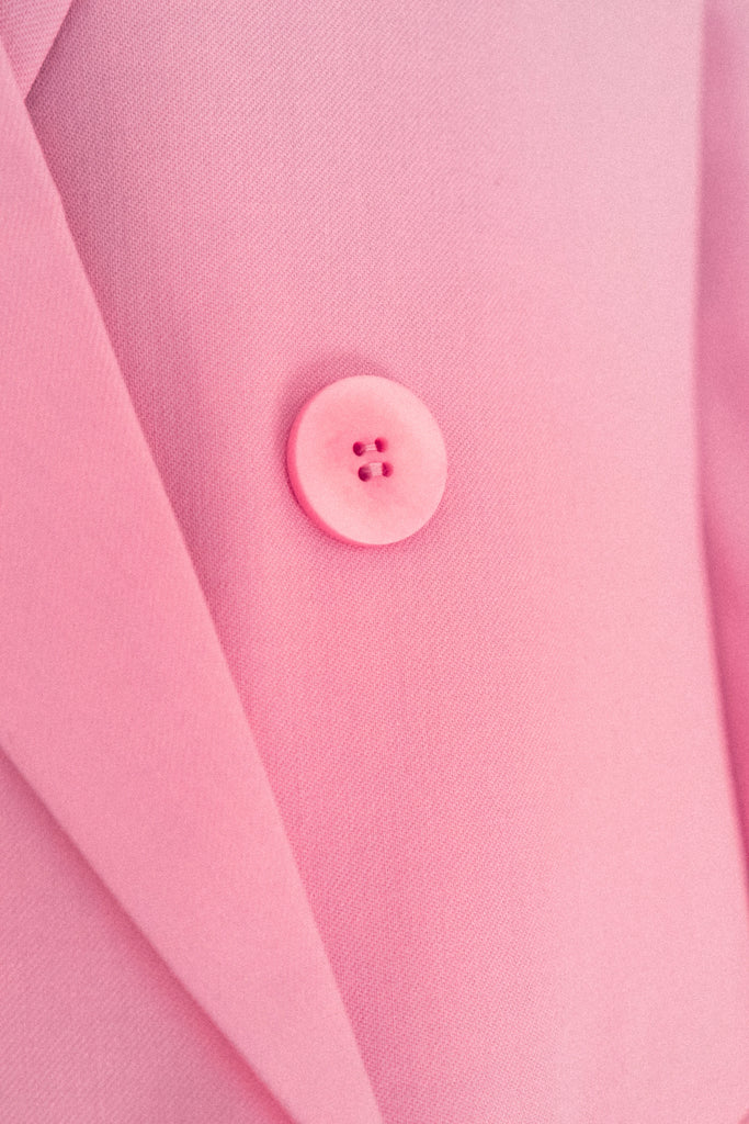 Close-up image of a pink button on the ALVA Blazer in Candy Pink. The button, featuring four holes, is prominently displayed on the left side and contrasts slightly with the fabric of the blazer. The material appears smooth, with the jacket lapel visible on the left side of the image.