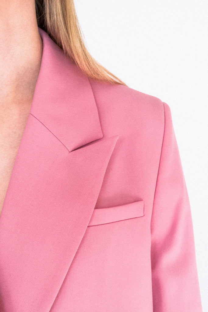 Close-up of a person wearing the ALVA Blazer in Candy Pink. Only a portion of the person’s face and shoulder are visible, along with the top half of the blazer, which features a notched lapel and a small chest pocket. The background is plain and light-colored.