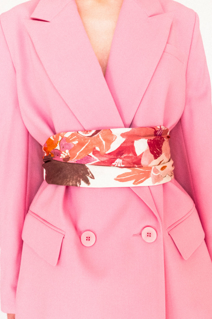 A person wearing the ALVA Blazer in Candy Pink, featuring large lapels and matching buttons, cinched at the waist with a wide, colorful floral-patterned scarf showcasing shades of red, pink, and beige. The background is plain white.