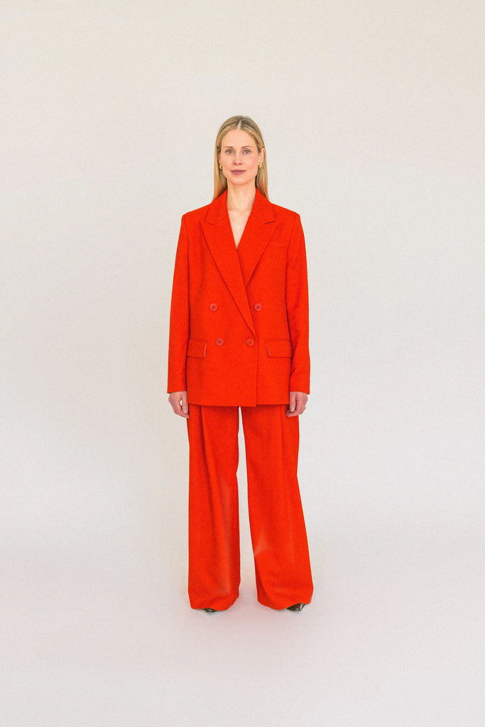A person with long blond hair stands against a plain white background, wearing the ALVA Blazer in Tangerine Red and matching pants. 
