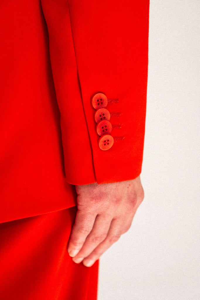 Close-up of a person wearing the ALVA Blazer in Tangerine Red. Only the sleeve with the buttons are visible. The fabric appears smooth and structured. The light background highlights the vibrant color of the blazer.