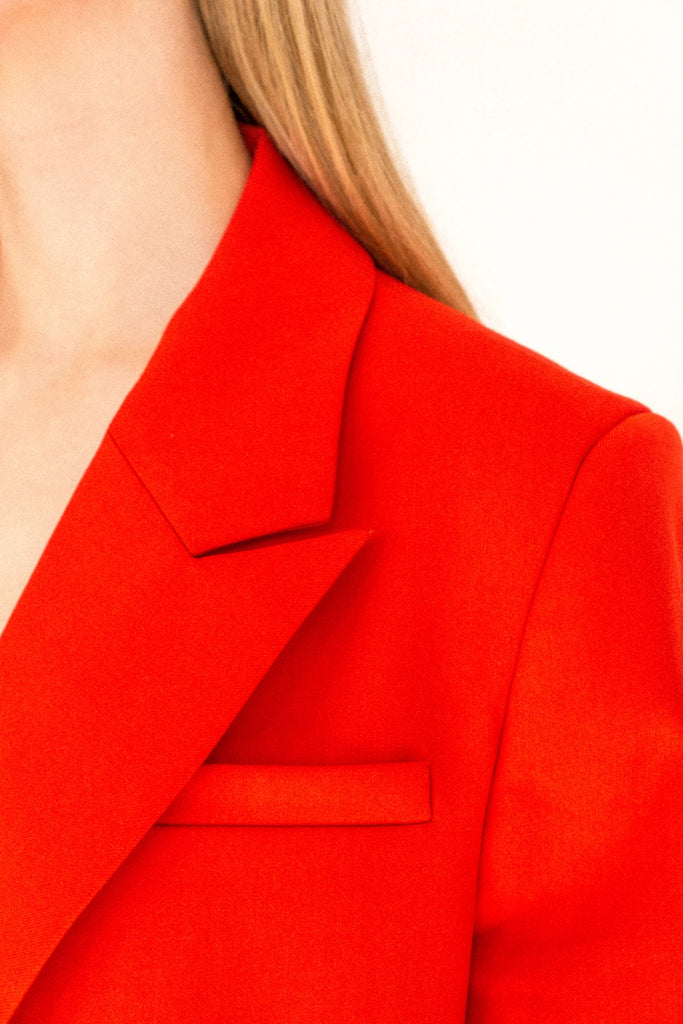 Close-up of a person wearing the ALVA Blazer in Tangerine Red. Only the shoulder, lapel, and part of the chest and collar are visible. The fabric appears smooth and structured, with a single pocket detail on the chest. The light background highlights the vibrant color of the blazer.