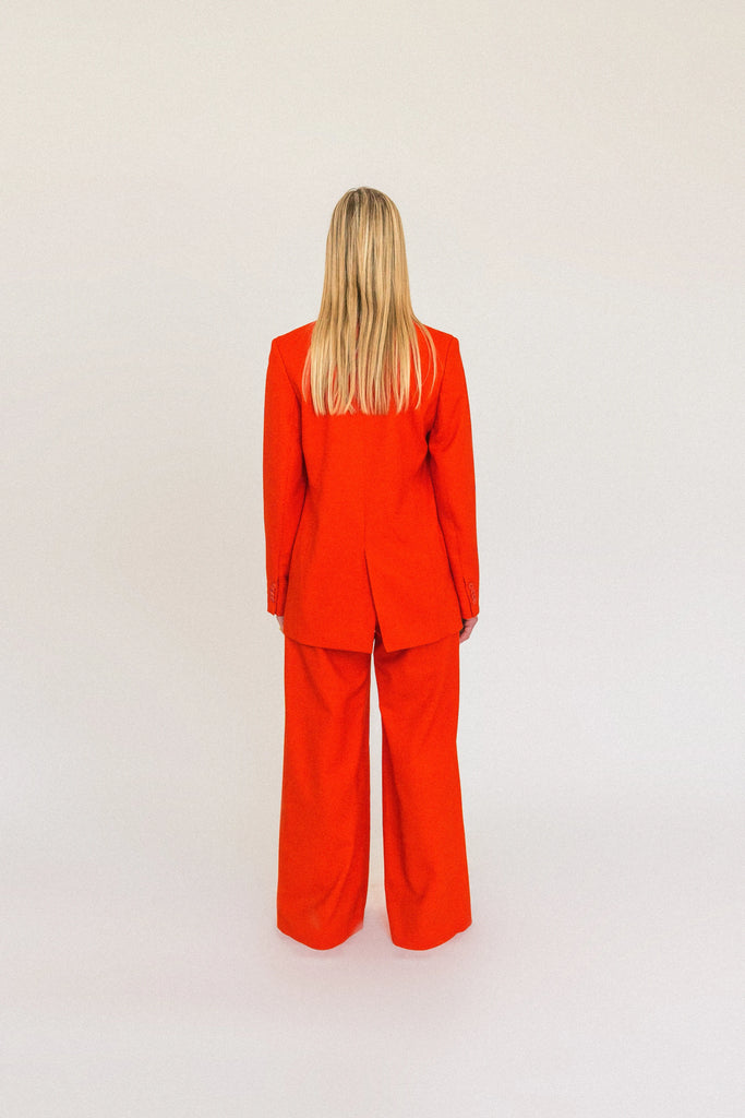 A person with long, blonde hair is standing facing away from the camera. They are wearing an oversized ALVA Blazer and matching pants, both in a vibrant tangerine red. The background is plain and light-colored.