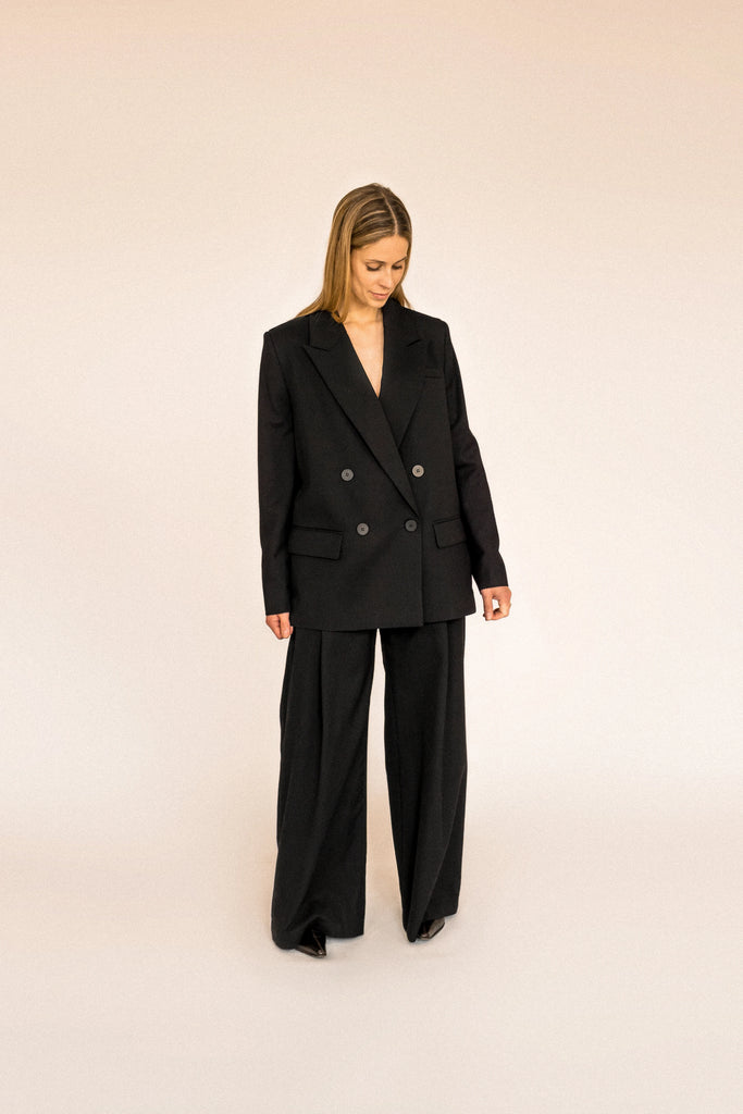A person with long blond hair stands against a plain white background, wearing the ALVA Blazer in Tuxedo Black and matching pants.