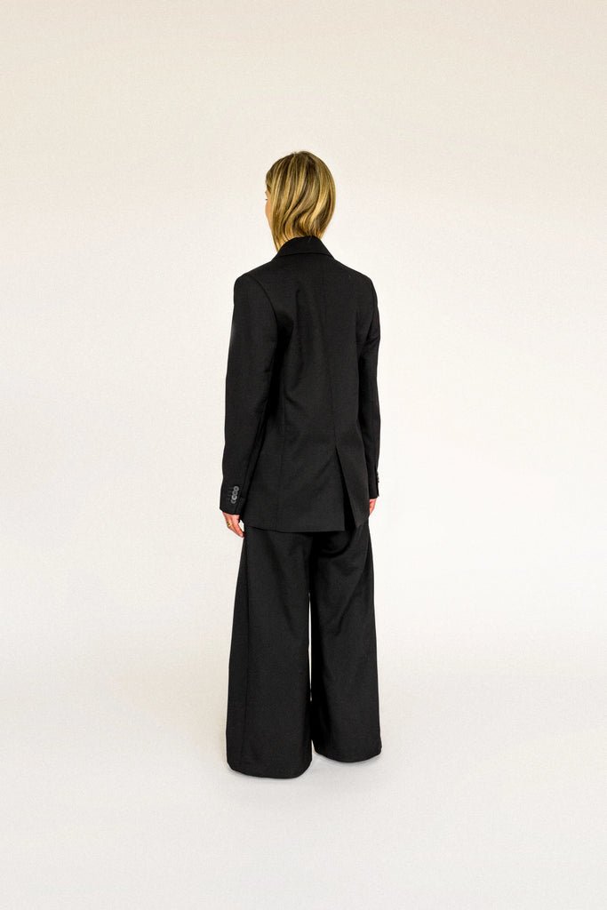 A person with long, blonde hair is standing facing away to the side. They are wearing an oversized ALVA Blazer and matching pants, both in a classic tuxedo black. The background is plain and light-colored.