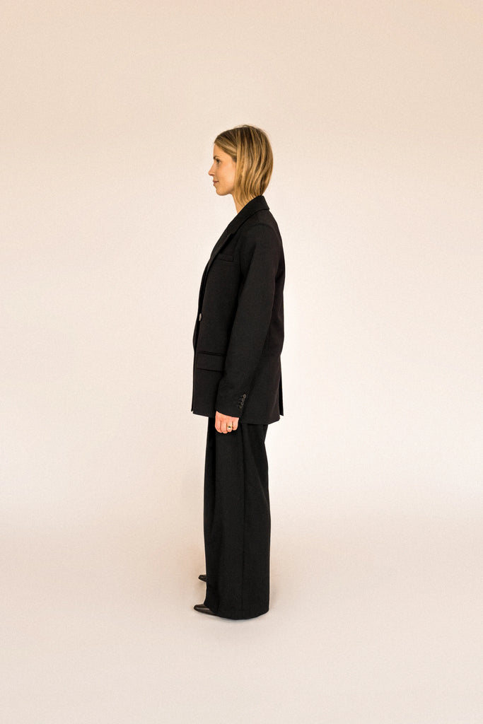 A person with long, blonde hair is standing facing away to the side. They are wearing an oversized ALVA Blazer and matching pants, both in a classic tuxedo black. The background is plain and light-colored.