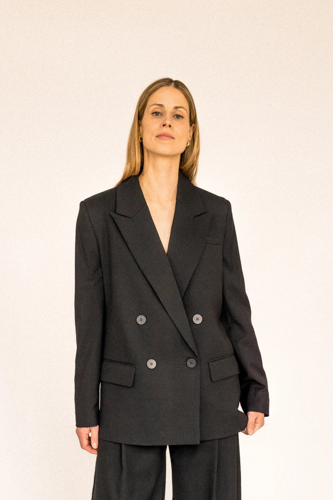 A person with long blond hair stands against a plain white background, wearing the ALVA Blazer in Tuxedo Black and matching pants. 