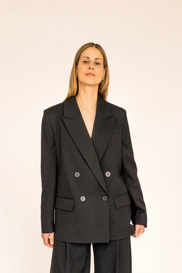 A person with long blond hair stands against a plain white background, wearing the ALVA Blazer in Tuxedo Black and matching pants. 