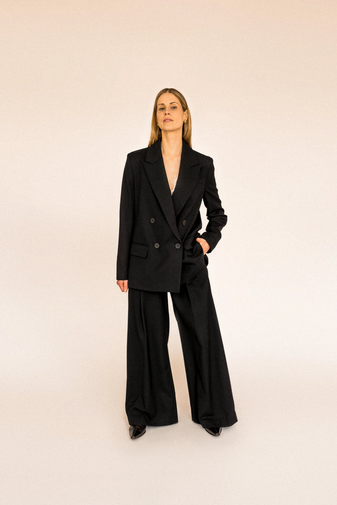 A person with long blond hair stands against a plain white background, wearing the ALVA Blazer in Tuxedo Black and matching pants.