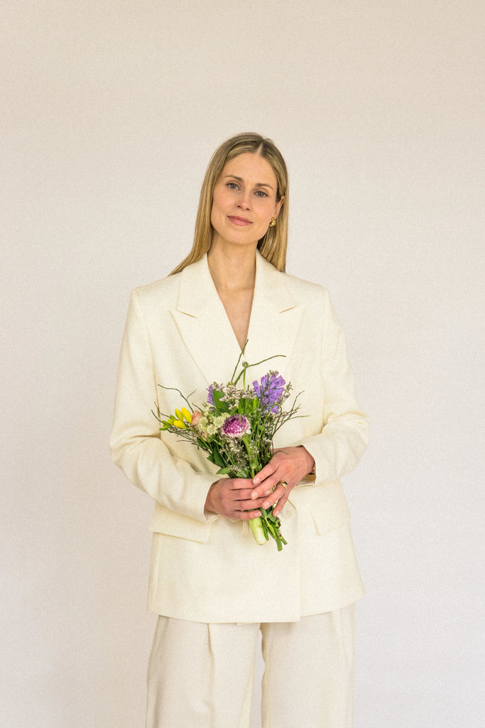 A person with long blonde hair is standing against a plain background, wearing an ALVA Blazer in Vanilla Creme. They are holding a small bouquet of colorful flowers and smiling gently at the camera.