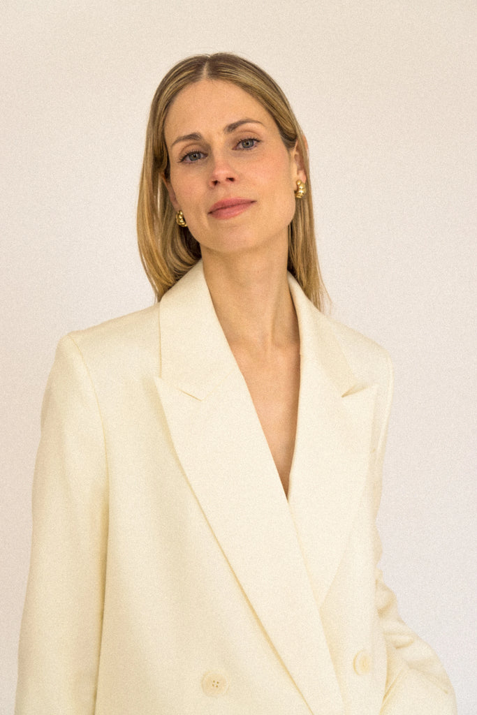 A person with long blond hair stands against a plain white background, wearing the ALVA Blazer in Vanilla Creme and matching shorts. Only the blazer is visible.