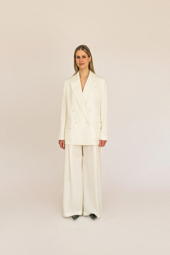 A person with long blond hair stands against a plain white background, wearing the ALVA Blazer in Vanilla Creme and matching pants.