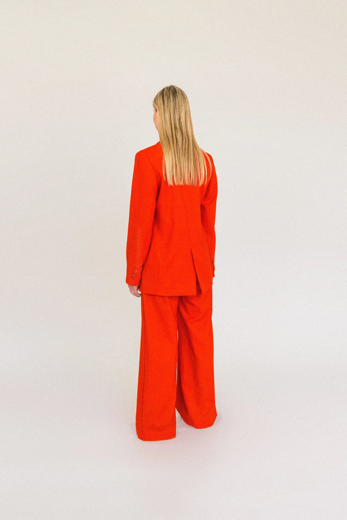 A person with long, blonde hair is standing facing away from the camera. They are wearing an oversized ALVA Blazer and matching pants, both in a vibrant tangerine red. The background is plain and light-colored.