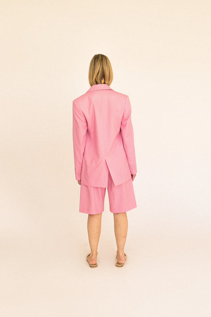 A person stands against a plain backdrop wearing the ALVA Blazer in Candy Pink, paired with matching knee-length shorts. They have shoulder-length blonde hair and are wearing beige slide sandals. Their expression is neutral, and they face the camera directly.