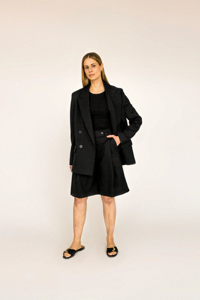 A person with long blond hair stands against a plain white background, wearing the ALVA Blazer in Tuxedo Black and matching shorts. 