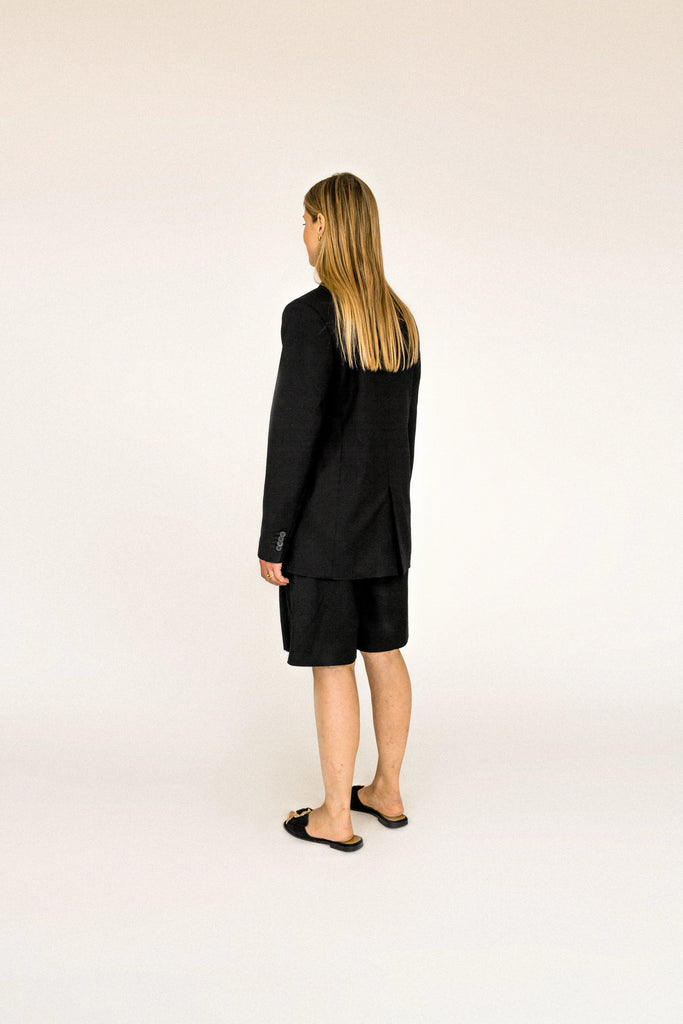 A person with long, blonde hair is standing facing away to the side. They are wearing an oversized ALVA Blazer and matching shorts.