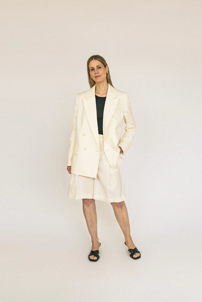 A person with long blond hair stands against a plain white background, wearing the ALVA Blazer in Vanilla Creme and matching shorts. 